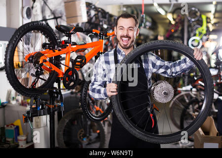 Sorridente uomo americano nel grembiule considerando la composizione della ruota per bicicletta Foto Stock