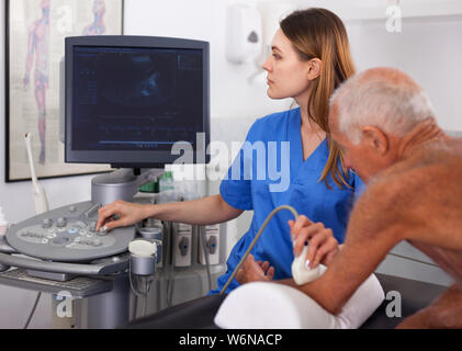 Diligente sorridente femmina piacevole il tecnico esegue la diagnosi paziente maschio utilizzando i moderni scanner ad ultrasuoni in clinica privata Foto Stock