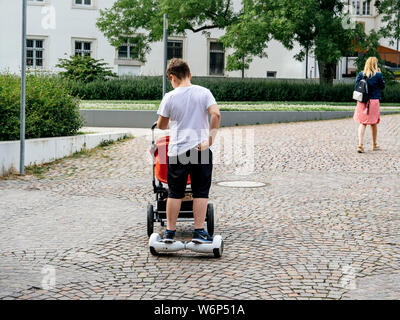 Baden-Baden, Germania - Luglio 7, 2019: vista posteriore del pigro giovane spingendo il carrello passeggino con il bambino mentre cavalcate un hoverboard elettrico sulle strade lastricate di Baden-Baden Foto Stock