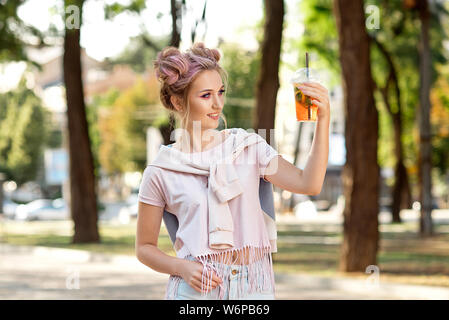 Giovane bella ragazza di bere succo di frutta fresco dalla plastica cibo da asporto tazze dopo una passeggiata all'aperto. Uno stile di vita sano. Slim sorridente bionda con capelli rosa Foto Stock