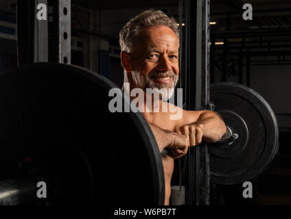 Ritratto di un affascinante vecchio atleta maschio con forti muscoli. L'atletico e sorridente uomo barbuto è appoggiata contro il barbell rack. Foto Stock