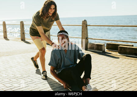 Giovane uomo essendo spinta su skateboard dal suo amico all'aperto su strada sul mare. Amici maschio avendo divertimento all'aperto su un giorno d'estate. Foto Stock