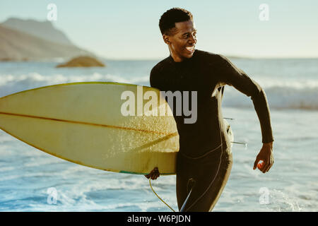 Sorridente maschio africana in esecuzione al di fuori dell'oceano dopo acqua surf. Felice giovane con le tavole da surf sulla spiaggia godendo le vacanze. Foto Stock