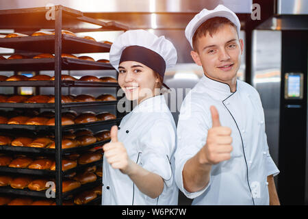 Giovane ragazzo e ragazza bakers sullo sfondo di un forno industriale in una panetteria mostrano il pollice verso l'alto. Foto Stock