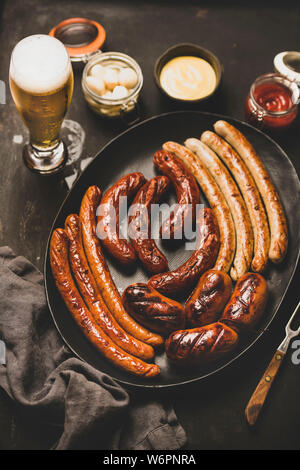 Oktoberfest dinner table concetto con grigliate di carne di vitello e di carne di maiale salsicce, la salsa in barattolo, decapati vaggies e birra in bicchieri su sfondo scuro, vista dall'alto Foto Stock