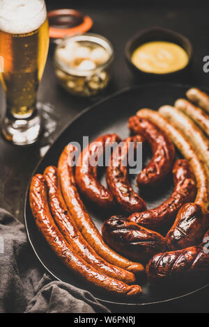 Oktoberfest dinner table concetto con grigliate di carne di vitello e di carne di maiale salsicce, la salsa in barattolo, decapati vaggies e birra in bicchieri su sfondo scuro, selectiv Foto Stock