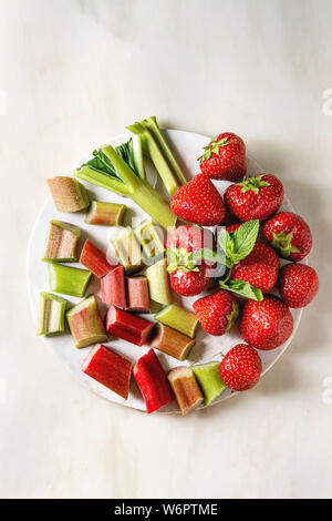 Fresco giardino organico di fragole e rabarbaro taglio steli sulla scheda di ceramica su marmo bianco dello sfondo. Appartamento laico, spazio. Ingredienti per l'estate lem Foto Stock