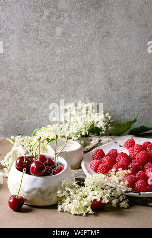 Freschi di ciliegia e frutti di bosco Lamponi nella tazza di ceramica e la piastra, elderflowers brocca di crema sul tavolo da cucina con parete grigia a sfondo. Spazio di copia Foto Stock