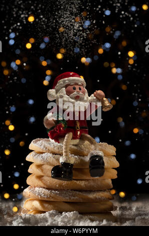 Babbo Natale seduto su una pila di ricco burro pezzata frollini con zucchero a velo con bokeh illuminazione effetto in background. Foto Stock