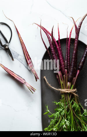 Un mazzetto di lavare le carote viola legati con spago di iuta su un carbone oblunga piatto. La superficie è una piastrella in marmo una carota dimezzata e una coppia di scisso Foto Stock