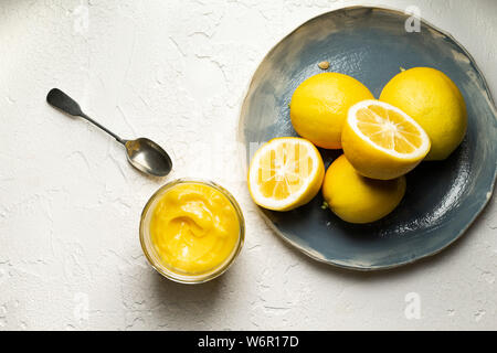 Limoni freschi interi e tagliate a metà, disposti su una mano blu piastra realizzata. Un vasetto di fatto in casa cagliata di limone e un cucchiaio sono a fianco. Foto Stock