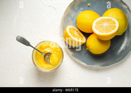 Limoni freschi interi e tagliate a metà, disposti su una mano blu piastra realizzata. Un cucchiaio nel vasetto di fatto in casa cagliata di limone è accanto a. Foto Stock