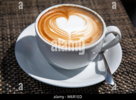 Tazza cappuccino con latte art cuore sulla parte superiore insieme al sole in un cafè all'aperto Foto Stock