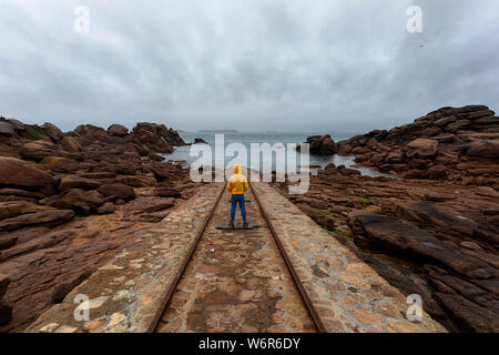 Francia, Bretagna, ragazzo in giacca gialla in piedi su rotaie in granito rosa di costa presso gli uomini Ruz faro di Ploumanach, guardando il mare Foto Stock