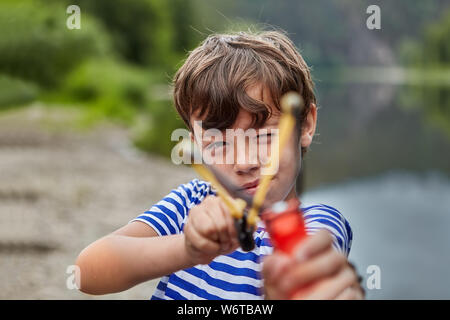 Little White Boy è in piedi nella foresta o pakr con slingshot nelle sue mani e la preparazione per la ripresa di spargimento fino ai suoi occhi, boy è il puntamento. Foto Stock