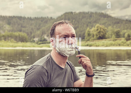Coppia uomo bianco è la rasatura il suo volto con rasoio sulla natura selvaggia accanto al fiume e alla foresta alla mattina, il suo volto è coperto dalla schiuma da barba, eco- Foto Stock