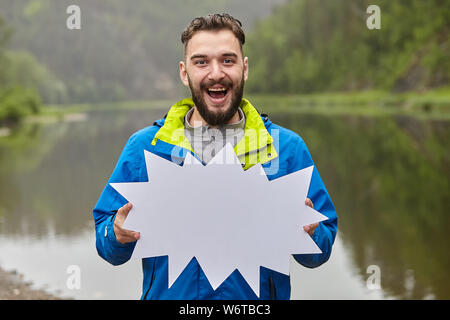 Giovane uomo caucasico con la barba è tenuta ripida vuoto di carta e sorridente, egli è nella foresta e vi è un fiume dietro di lui, il turismo ecologico. Foto Stock