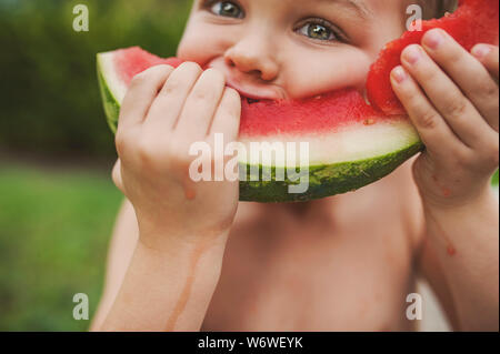 Un giovane bambino sta assumendo un boccone fuori di una grande fetta di anguria e guardando direttamente nella fotocamera. Foto Stock