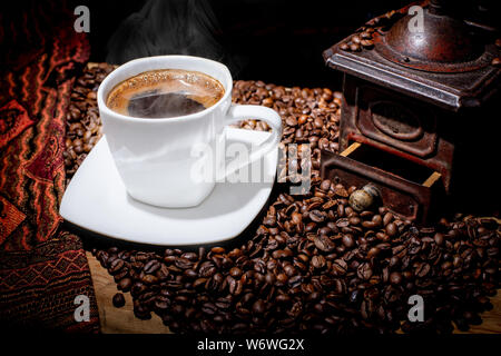 White tazza di caffe' fumante. Un tavolo di legno ricoperto con semi tostati.. Vintage, bronzo retrò coffee mill su sfondo nero. Rosolare i chicchi di caffè. C Foto Stock