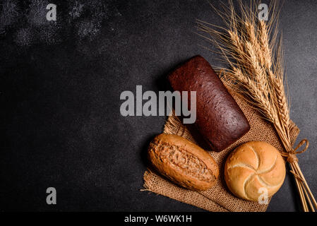 Fresco e fragrante pane con grani e coni di grano contro uno sfondo scuro. Assortimento di pane cotto sul tavolo di legno sfondo Foto Stock