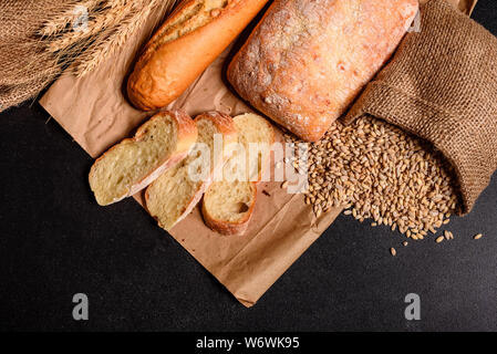 Fresco e fragrante pane con grani e coni di grano contro uno sfondo scuro. Assortimento di pane cotto sul tavolo di legno sfondo Foto Stock