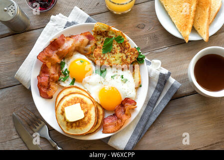 Una colazione americana completa su legno, vista dall'alto. Sunny Side uova fritte, pancetta arrosto, hash brown, frittelle, toast, succo d'arancia e caffè per la prima colazione Foto Stock