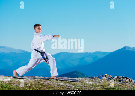 Il Karate uomo in un kimono esegue una mano anteriore kick (Choku-zuki) mentre si sta in piedi sul prato verde sulla cima di una montagna. Foto Stock