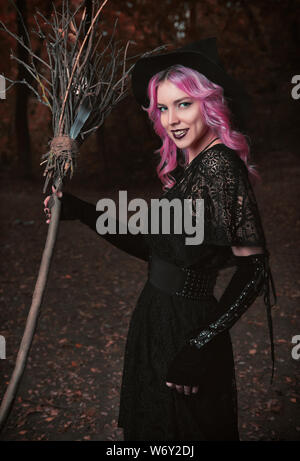 Tema Halloween: contemporanea sorridente giovane strega vestito in abito nero e il cappello con la scopa in una foresta scura. Tema Halloween Foto Stock