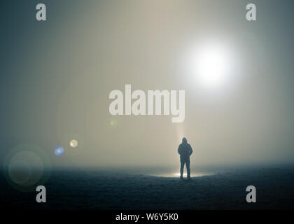 Un lone incappucciati figura in piedi in un campo su un losco, notte di nebbia con UFOsfloating nel cielo. Foto Stock