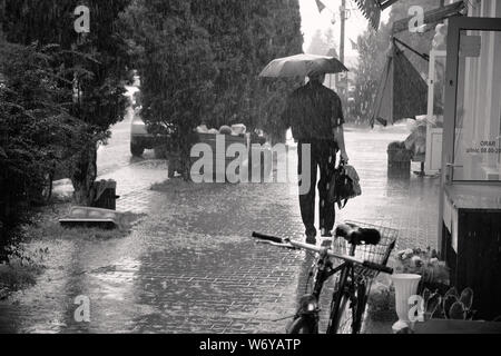 Uomo con valigetta a piedi sotto la pioggia con ombrello. Pioggia creando enormi pozzanghere sul marciapiede Foto Stock