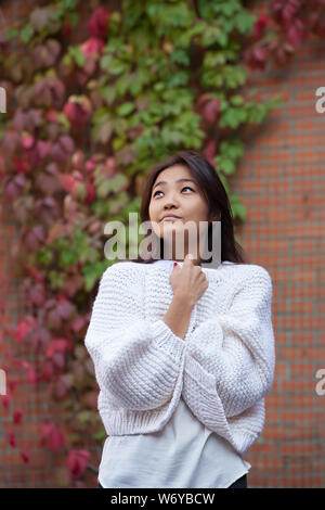 Bella asiatica giovane donna guarda.ella è in maglia bianco cardigan vicino a un muro di mattoni e riccio wild multi-colore di uva Foto Stock