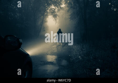 Una solitaria, incappucciati figura in piedi di fronte a una macchina guardando un vuoto misty country road profilarsi di notte in auto fari anteriori Foto Stock