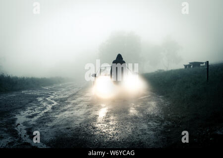 Una solitaria, Spooky, incappucciati figura in piedi di fronte a una macchina guardando un vuoto misty country road profilarsi di notte in auto fari. Foto Stock