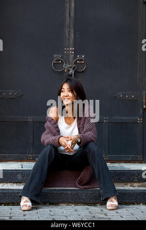 Bella ragazza asiatica in un viola cardigan lavorato a maglia si siede sulle scale vicino a casa Foto Stock