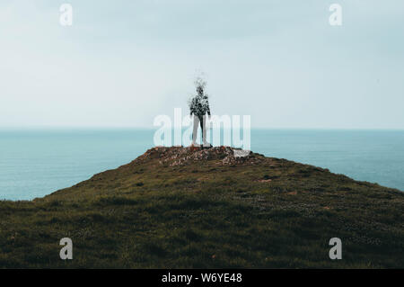 Un lone incappucciati figura permanente sulla cima di una collina e di disintegrazione guardando fuori attraverso l'oceano. Foto Stock