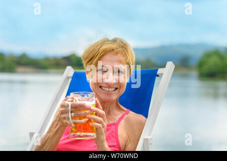 Felice donna gentile con un caldo sorriso accogliente che gode di una grande il boccale di birra fredda come lei si rilassa in un lago su vacanze estive Foto Stock
