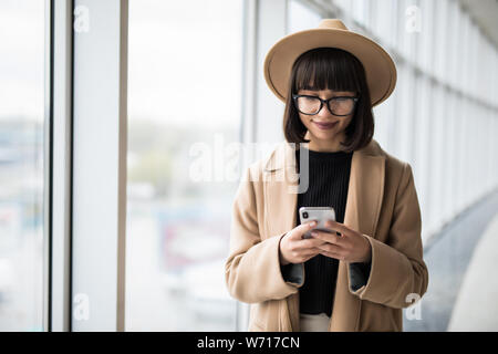 Giovani orgogliosi businesswomanwear in cappotto e hat utilizzare il telefono cellulare mentre si è in piedi vicino alla finestra Foto Stock