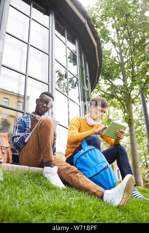 Angolo basso ritratto di due studenti seduti sul prato verde nel campus e studiando all'aperto, spazio di copia Foto Stock