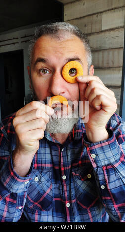 Adulto felice uomo barbuto detiene due biscotti a forma di ciambella e un mese davanti al suo volto, raffigura un monocolo e baffi, concetto di divertimento con c Foto Stock