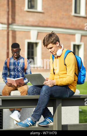 A piena lunghezza ritratto contemporaneo di studente di college utilizzando laptop seduto sul tavolo all'aperto in campus, spazio di copia Foto Stock