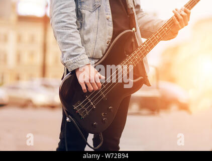 Street giovane musicista, vestito con una camicia di denim e illuminata dalla luce del sole, giocando a basso elettrico chitarra potente via Foto Stock