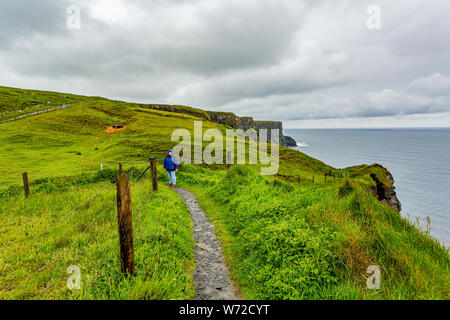 Donna in giacca blu godendo la passeggiata costiera percorso da Doolin per le scogliere di Moher, geosites e geoparco, Wild Atlantic modo la molla Rainy day Foto Stock