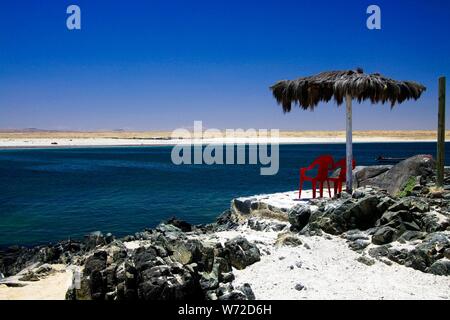 Spiaggia di sabbia bianca (Playa blanca) sulla costa pacifica del deserto di Atacama con sedie isolate di plastica rossa sotto ombrello con tetto di paglia Foto Stock