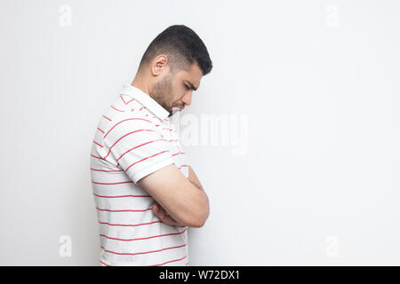 Profilo vista laterale verticale della sola SAD o premuto barbuto giovane striped t-shirt in piedi tenendo la sua testa in basso e la sensazione di cattivo o di pensare. Foto Stock