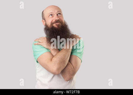 Ritratto di smiley e sognanti di mezza età uomo calvo con barba lunga in verde t-shirt standing, abbracciando stesso, che guarda lontano e sognare qualcosa. Foto Stock