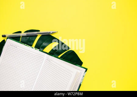 Il Blocco note di bianco e una penna per scrivere, monstera foglio su sfondo giallo, copia dello spazio. Foto Stock