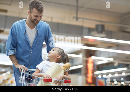 Ritratto di felice giovane padre a fare la spesa nel supermercato e sorridente a poco ragazza seduta nel carrello, spazio di copia Foto Stock