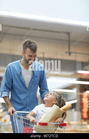 Ritratto di felice giovane padre a fare la spesa nel supermercato e sorridente a poco ragazza seduta nel carrello Foto Stock