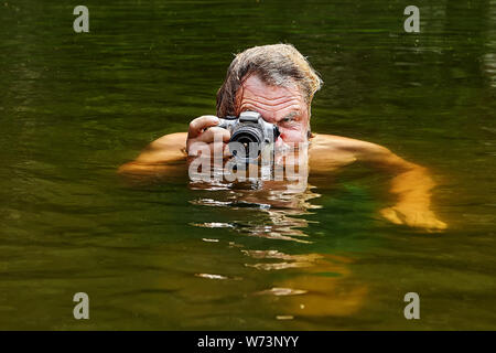 Uomo maturo è nuotare nel fiume e scattare foto tenere la fotocamera sulla superficie dell'acqua, ecoturismo. Foto Stock