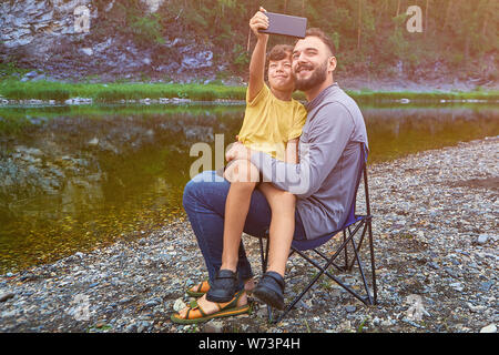 Giovani allegro uomo caucasico e il suo piccolo figlio vengono scattate le foto sul litorale vicino al fiume, essi sono seduti sulla sedia e sorridente. Foto Stock